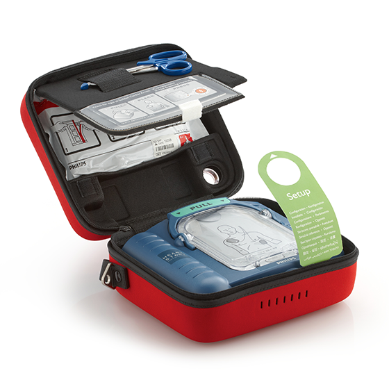 The red first aid kit bag is open, containing medical scissors, supplies, and instructions with a setup guide tag hanging out, alongside the Philips HeartStart OnSite Defibrillator - Complete Package by Philips.