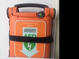 Orange Powerheart G5 AED (Automated External Defibrillator) mounted on a white wall, secured with a black strap and an adjustable depth Universal AED Bracket from the AED Shop, crafted from rugged steel.